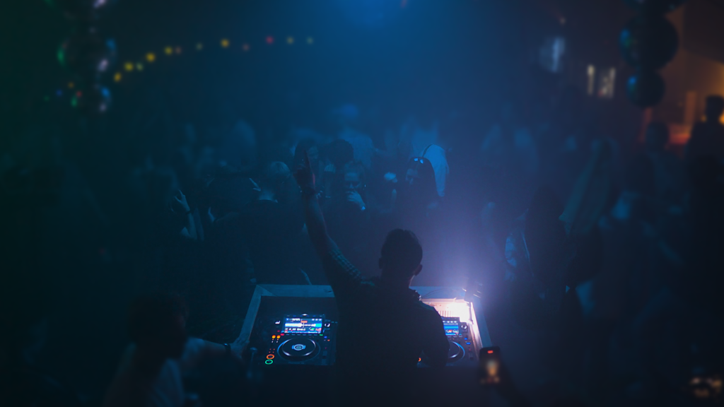 R-TLN performing a live DJ set, mixing energetic Trance and Hard House tracks, with colorful lights and an enthusiastic crowd in the background.