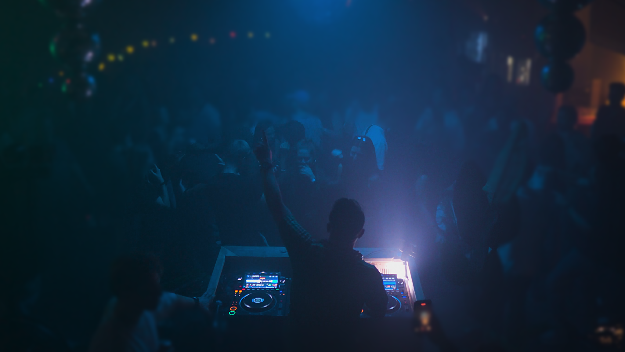 R-TLN performing a live DJ set, mixing energetic Trance and Hard House tracks, with colorful lights and an enthusiastic crowd in the background.