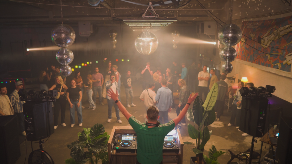 R-TLN performing a live DJ set, mixing energetic Trance and Hard House tracks, with colorful lights and an enthusiastic crowd in the background.